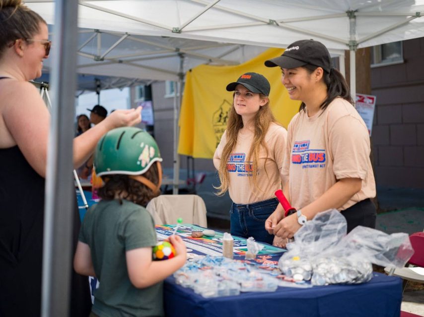 At an outdoor booth filled with merchandise and promotional items, two women assist visitors, including a child wearing a helmet modeled after the iconic design of Prop L.