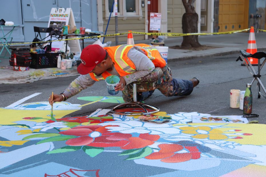 A person wearing safety gear paints a colorful street mural with flowers. Painting supplies are scattered nearby, and traffic cones are set up around them.