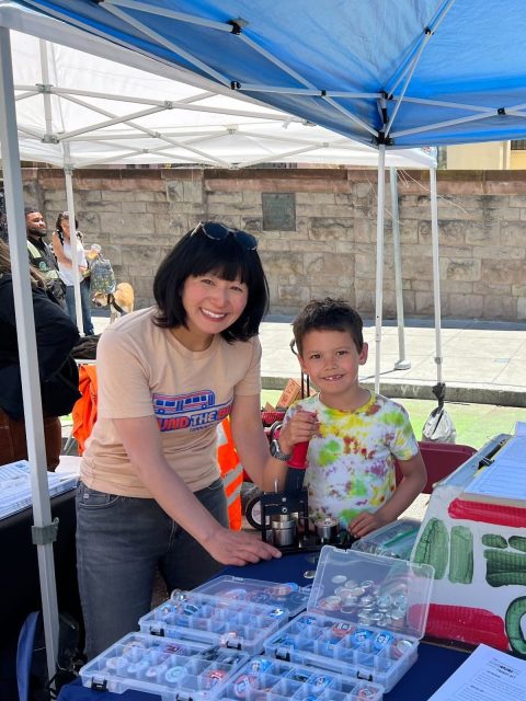 A woman and a child smile at a booth filled with button-making materials, the perfect prop for their creative afternoon. They stand cheerfully under a canopy outdoors.