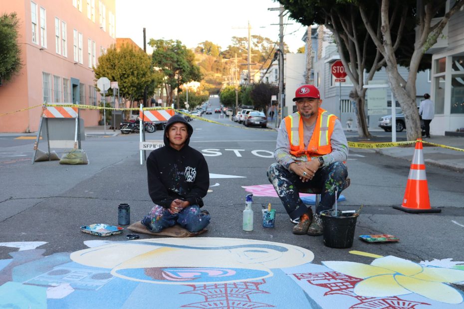 Two artists sitting on a street painting a mural, surrounded by traffic cones and barriers.