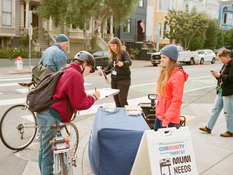 People gathering at a table on a sidewalk, with one person signing a paper and another holding a tablet, surrounded by bicycles and a sign advocating Prop L.