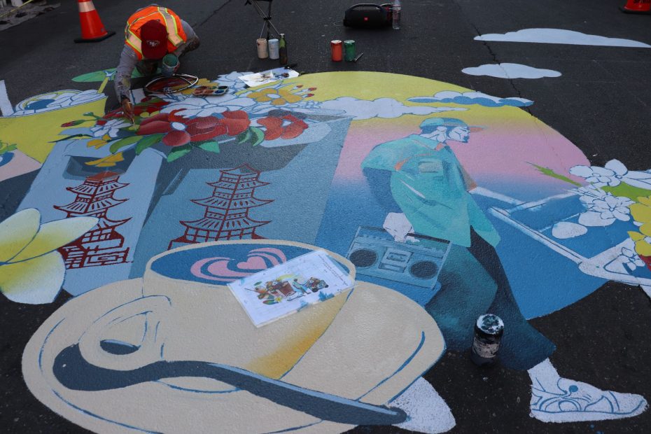 Artist painting a colorful street mural featuring a person with headphones, flowers, pagodas, a boombox, and a teacup, with traffic cones and paint supplies nearby.
