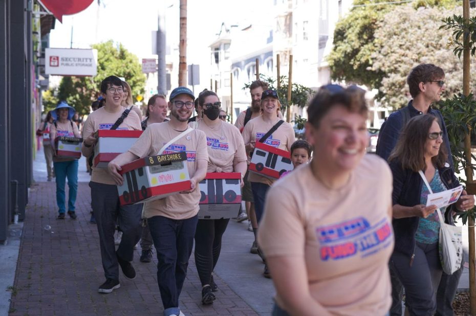 A group of people wearing matching shirts strolls down the sidewalk, carrying prop boxes decorated to resemble buses.
