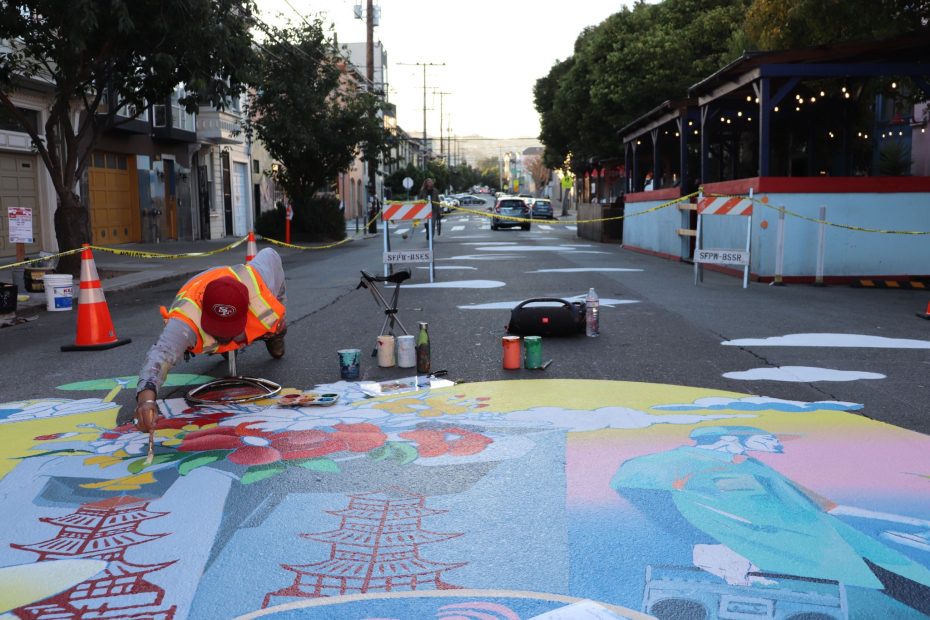 Artist painting a colorful mural on a closed-off street, surrounded by safety cones and barriers.