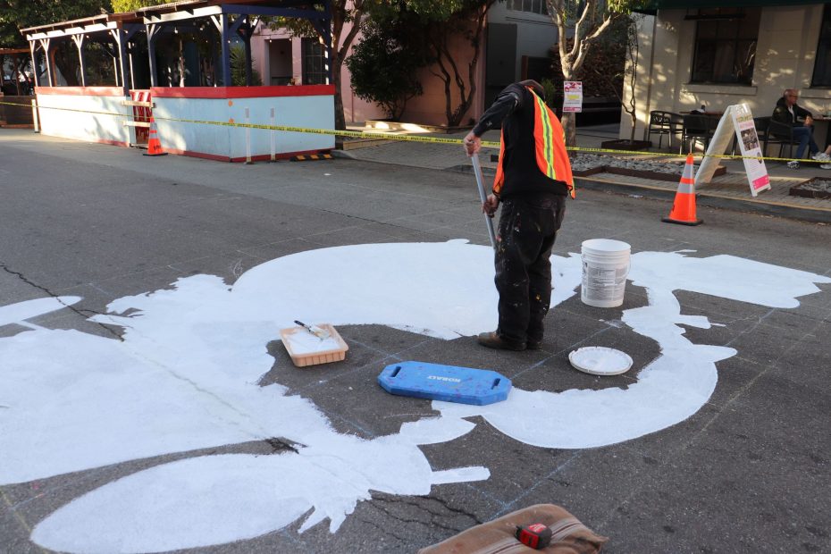 Person in a safety vest painting a large white shape on a street. Traffic cones and barriers are visible around the area.