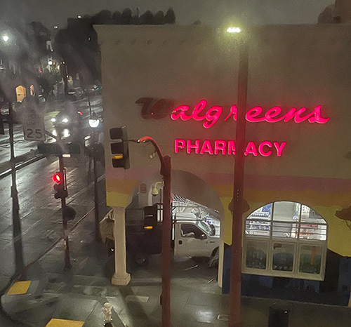 Night street view of a Walgreens Pharmacy with illuminated signage, traffic lights, and a few vehicles on a wet road.