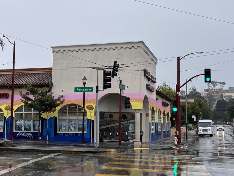 Street corner with a colorful mural on a building, wet pavement, and a traffic light. A white truck is stopped at the intersection on a rainy day.