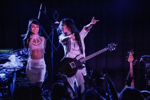 Two performers on stage; one holds a guitar while gesturing, and the other stands beside. The scene is dimly lit with musical equipment visible in the background.