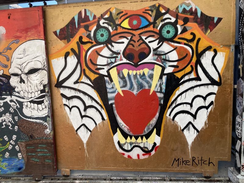 Colorful graffiti art of a tiger with exaggerated features, including a heart-shaped nose and wide eyes, on a wooden panel. Other artwork is partially visible on either side.