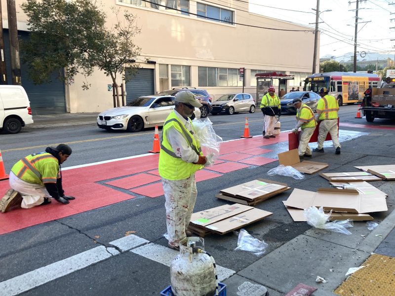 Workers in reflective vests paint red bus lanes on a city street, with equipment and cardboard on the ground. Traffic is passing by.