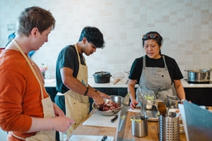 Three people in a kitchen cooking. One prepares food in a bowl while the others watch. Various cooking utensils and pots are visible on the counter.