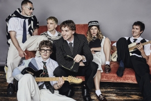 A group of six people sitting on and around a red couch, holding musical instruments. They are dressed in casual, eclectic clothing against a plain backdrop.