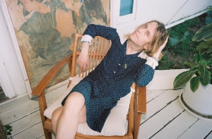 Person with light hair sitting on a wooden chair outdoors, wearing a dark polka dot dress, with a relaxed posture next to a potted plant.