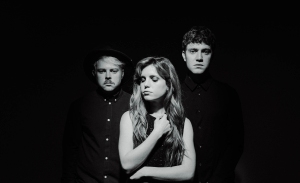 Three people are standing against a dark background, wearing dark clothing. The person in front has long hair and closed eyes, while the two behind have short hair and are looking forward.