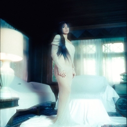 A woman in a long, light-colored dress stands in a dimly lit room with a covered armchair and a lamp on a table.