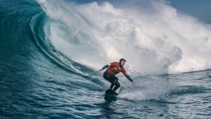 A surfer in a red wetsuit rides a large wave in the ocean under a partly cloudy sky.