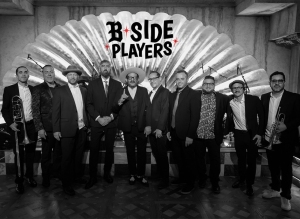 A group of ten men, some holding musical instruments, stand together in front of a large decorative shell backdrop with "B Side Players" written above them.