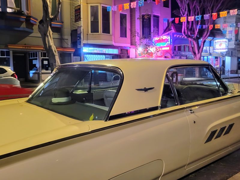 A vintage yellow car is parked on a street at night, with lit storefronts and colorful flags overhead.