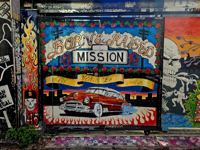 Graffiti mural featuring a classic car with the text "Born & Raised Mission," surrounded by colorful designs, skulls, and various tags.