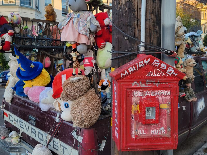 A truck with stuffed animals attached to its back, parked on the street beside a red fire alarm box.