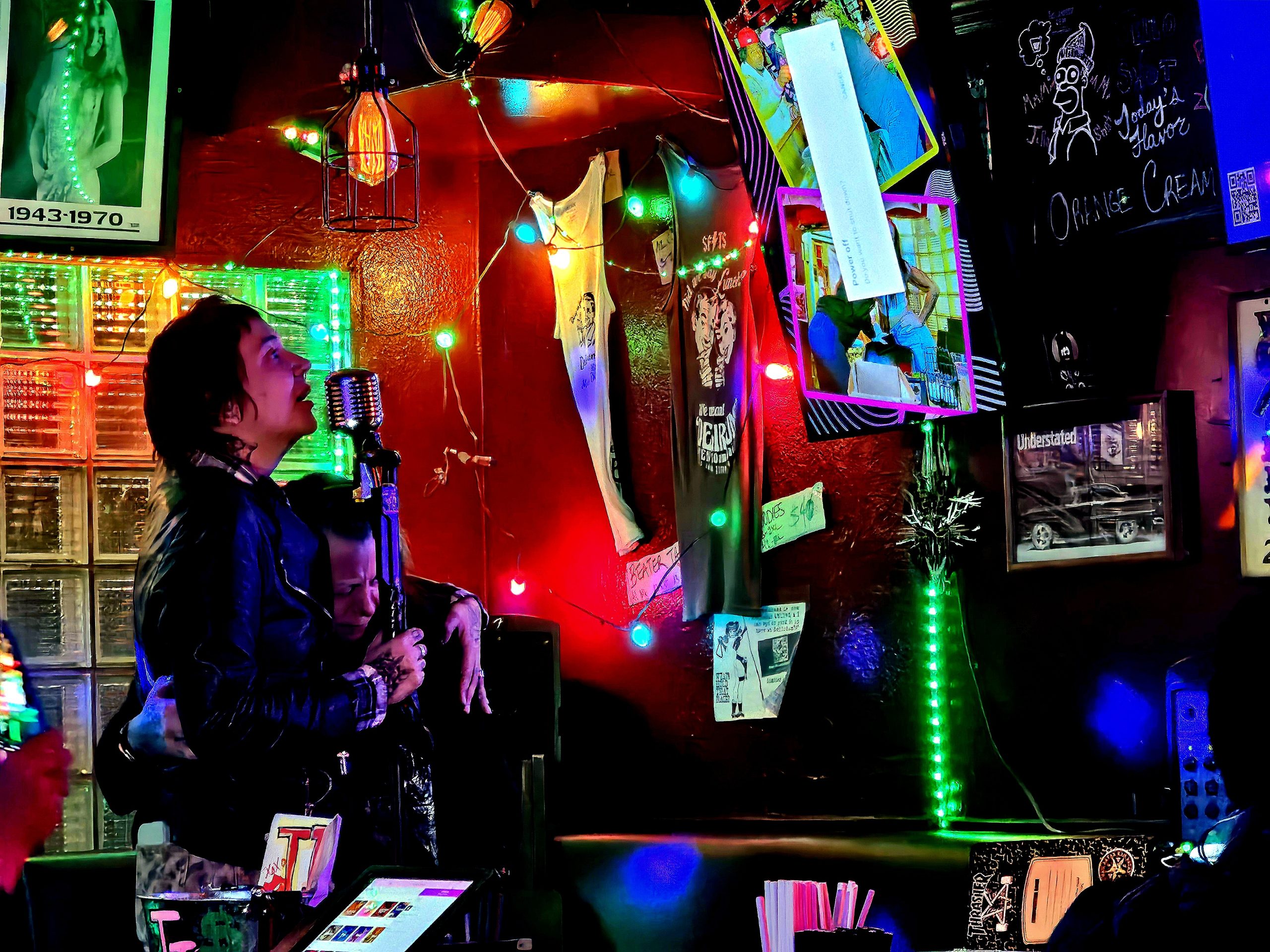 Person singing into a microphone in a dimly lit room decorated with colorful lights and posters.