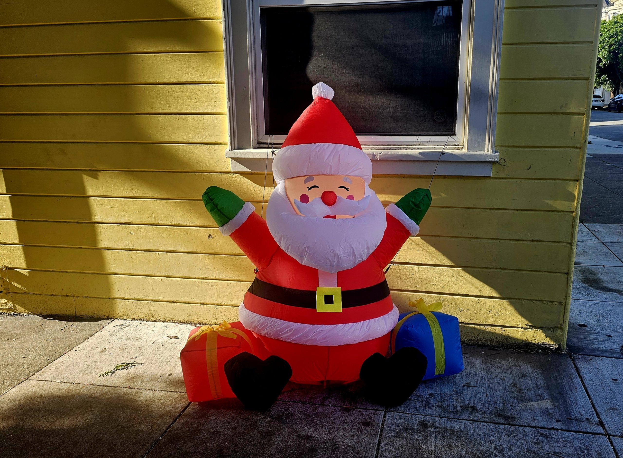 Inflatable Santa Claus decoration sits on a sidewalk beside a yellow building, with two gift boxes, one red and one blue.