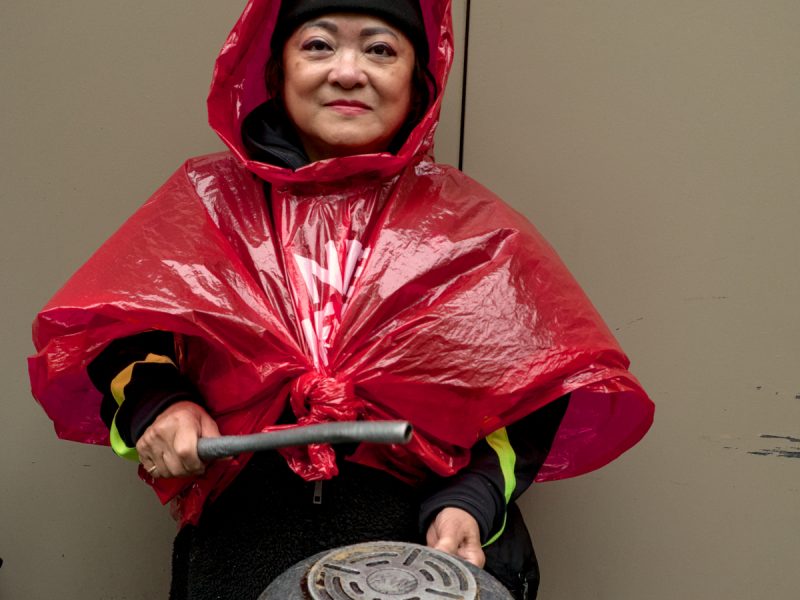 Person in a red poncho and beanie holds a metal pot and stick, standing against a plain wall.