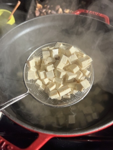 A metal strainer holds small cubes of tofu above a steaming pot of water.