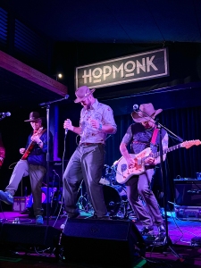 Three musicians perform on stage at HopMonk, with two playing guitars and one singing. They wear casual clothing, and cowboy hats under purple lighting.