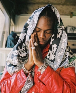 Person wearing a patterned headscarf and red jacket, with eyes closed and hands in a prayer gesture, stands indoors.