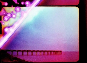 A vintage-style photo shows a pier extending into the sea beneath a colorful, psychedelic sky with pink and purple hues.