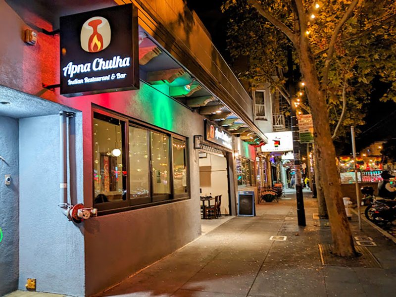 Streetview of "Apna Chulha," an Indian restaurant and bar, with festive lights and a sidewalk lined with trees.