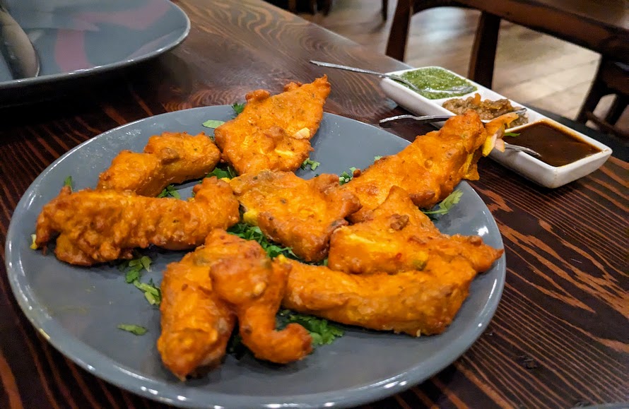 Plate of fried fish pieces on a dark plate with two small bowls of green and brown sauces on a wooden table.