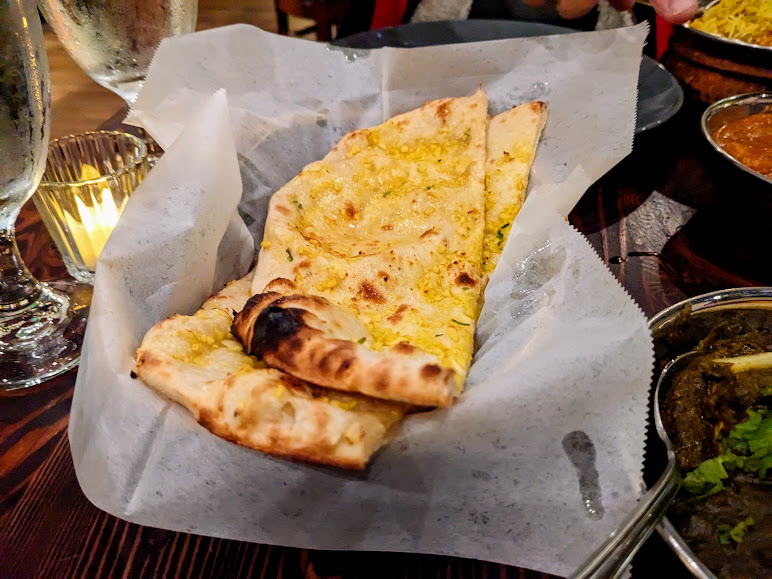 Basket of garlic naan on parchment paper, placed on a wooden table beside glasses and a dish with sauce.