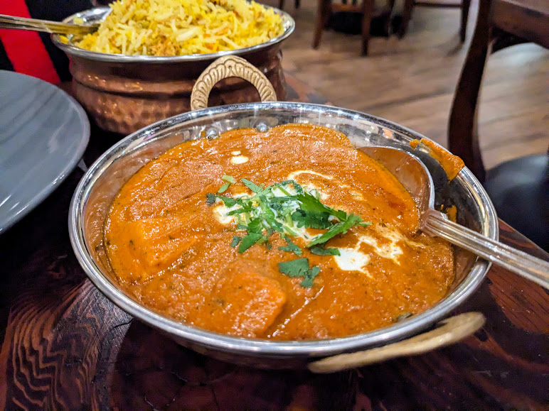 A bowl of curry garnished with cilantro sits on a wooden table, accompanied by a dish of biryani.