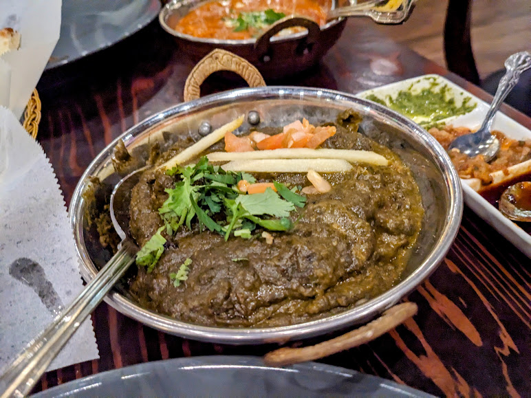 A plate of saag curry garnished with ginger and cilantro, alongside a side of sauces, on a dark wooden table.