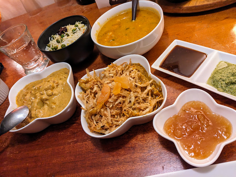 A spread of Indian dishes on a wooden table, featuring rice, curry, dal, and assorted sauces in white and black bowls, with a glass of water.