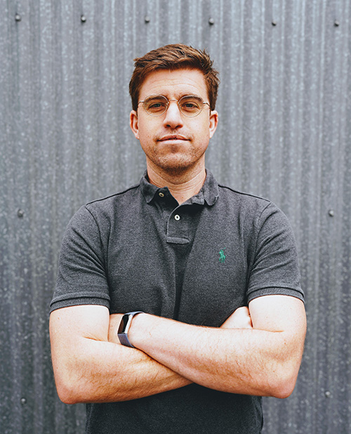 A man in a dark polo shirt stands with arms crossed against a corrugated metal background.