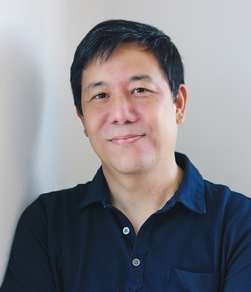 A person with short dark hair, wearing a dark blue collared shirt, stands against a light-colored wall, arms crossed and smiling slightly.