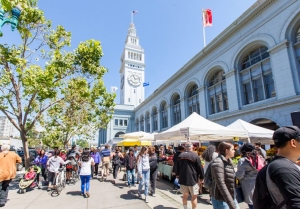 People walk by the Ferry Building Marketplace with vendor tents and a clock tower in view.