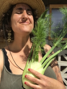 Person smiling while holding a fennel bulb, wearing a straw hat and earrings.