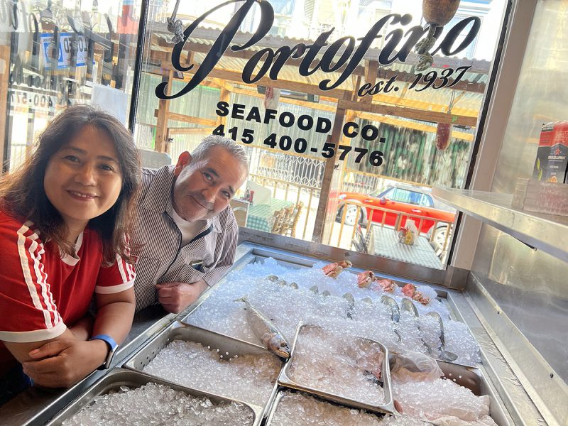 Two people stand next to a seafood display on ice inside Portofino Seafood Co. with a window showcasing the street outside.