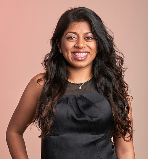 Smiling person with long hair, wearing a sleeveless black top, stands against a neutral background.