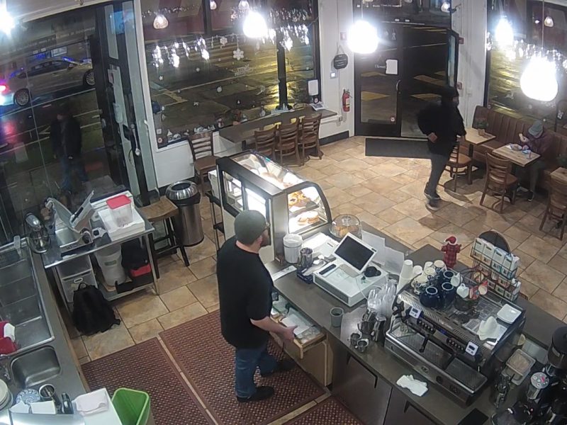 A cafe interior with a staff member behind the counter and a customer approaching, unaware of an attempted robbery that shook the cozy space. The brightly lit room features tables against the walls and display cases showcasing pastries, adding a deceptive calm to the air. .