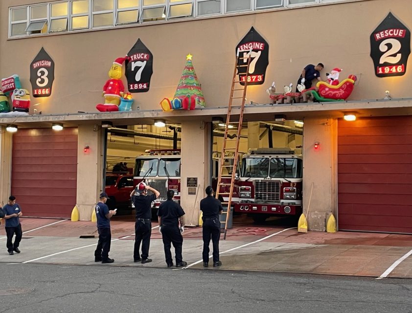 Firefighters decorate a fire station with inflatable holiday figures above garage doors, while two fire trucks are parked inside.