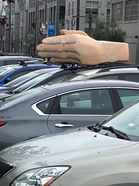 A large hand sculpture is mounted on top of a gray car parked in a parking lot.