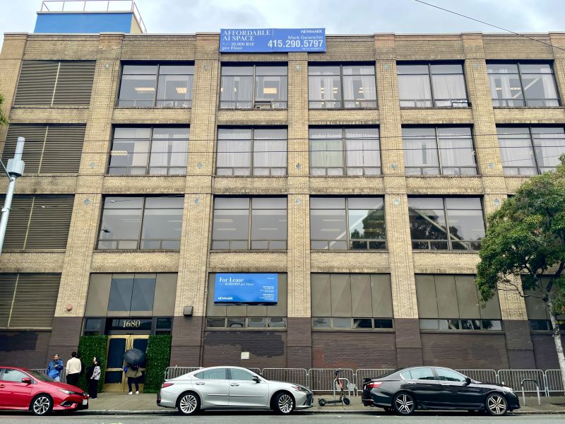 A multi-story brick building with "For Lease" sign, ground-level people and cars parked on the street in front.