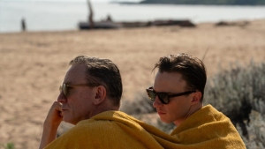 Two people wrapped in yellow towels are sitting on a sandy beach with sunglasses, looking towards the sea.