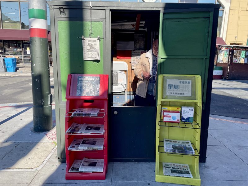 A person stands inside a small kiosk selling newspapers. Two racks outside display publications, one red and one yellow. The kiosk is situated on a sidewalk in a sunny area.