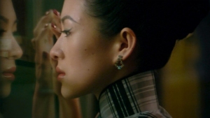 A woman in profile with a high collar and earrings gazes at her reflection in a glass surface.
